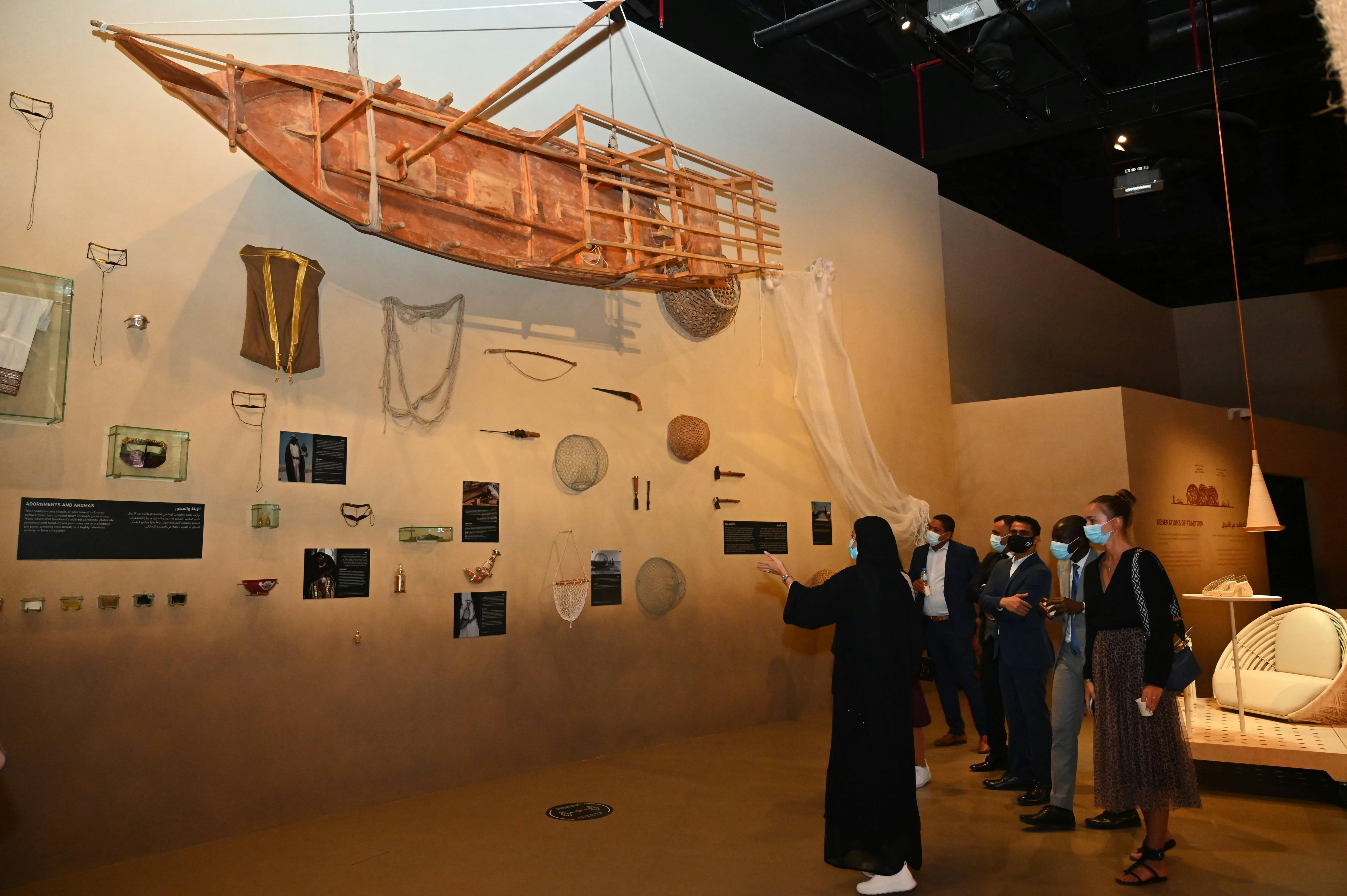 Museum exhibit with various artifacts on a wall, including a suspended wooden boat. A person in black points at the display while others watch.