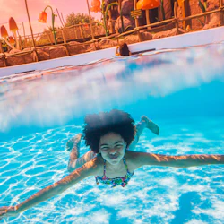 A child swimming underwater in a pool, with a theme park featuring large decorative mushrooms visible above water.