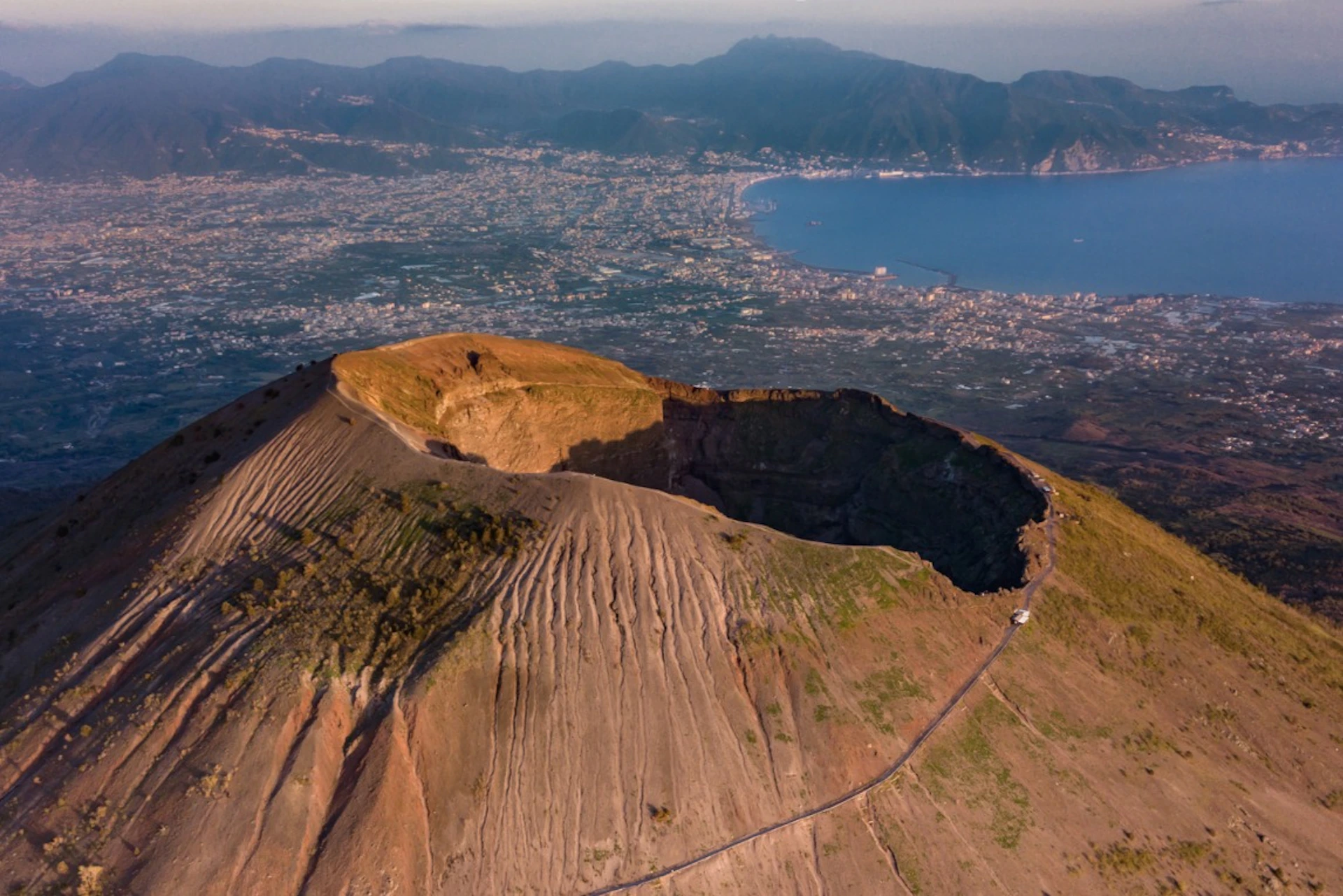 An aerial view of a volcanic crater on a mountain with a city, coastline, and mountains in the background.