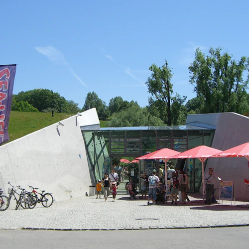 Menschen versammeln sich unter roten Schirmen vor einem Gebäude mit der Aufschrift "SEA LIFE", in dessen Nähe Fahrräder geparkt sind. Grünzeug und Fahnen sind zu sehen.