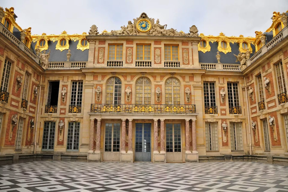 Facade of an ornate historical building with decorative details, large windows, and a checkered courtyard in front.