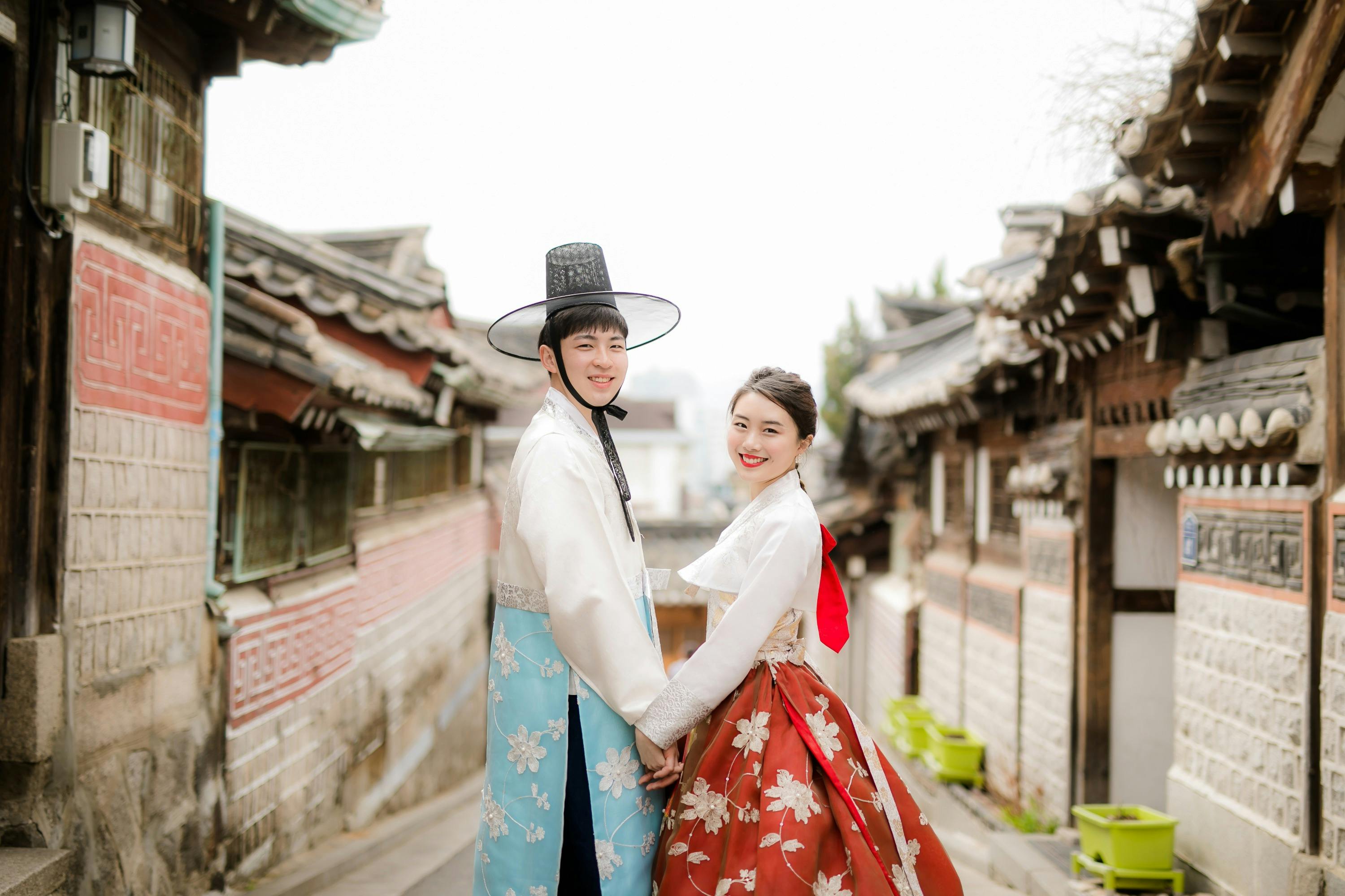 A couple in traditional Korean attire poses outdoors in a historic neighborhood with tiled roofs and narrow pathways.