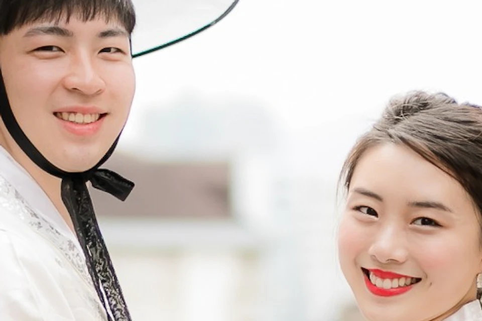 A couple wearing traditional Korean hanbok standing in a narrow street lined with traditional buildings, holding hands and smiling.