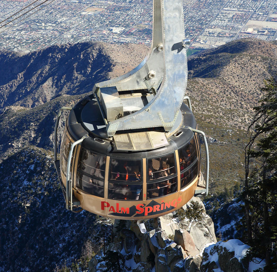 Aerial tramway car labeled "Palm Springs" ascending over mountainous terrain with a cityscape in the background.