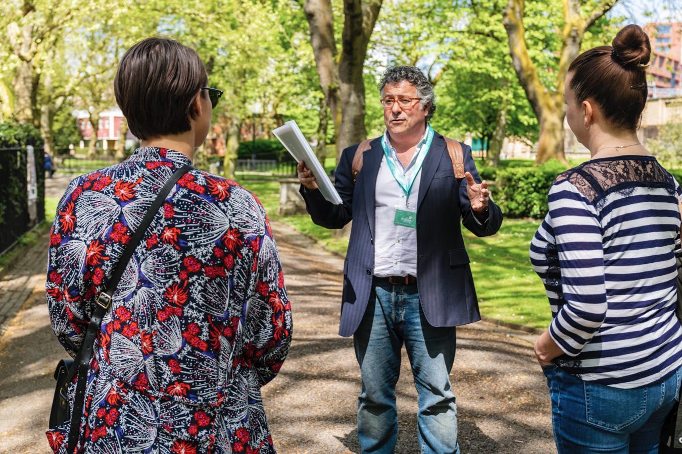 A man with a folder gestures while talking to two women in a park with trees and a sunny, green background.