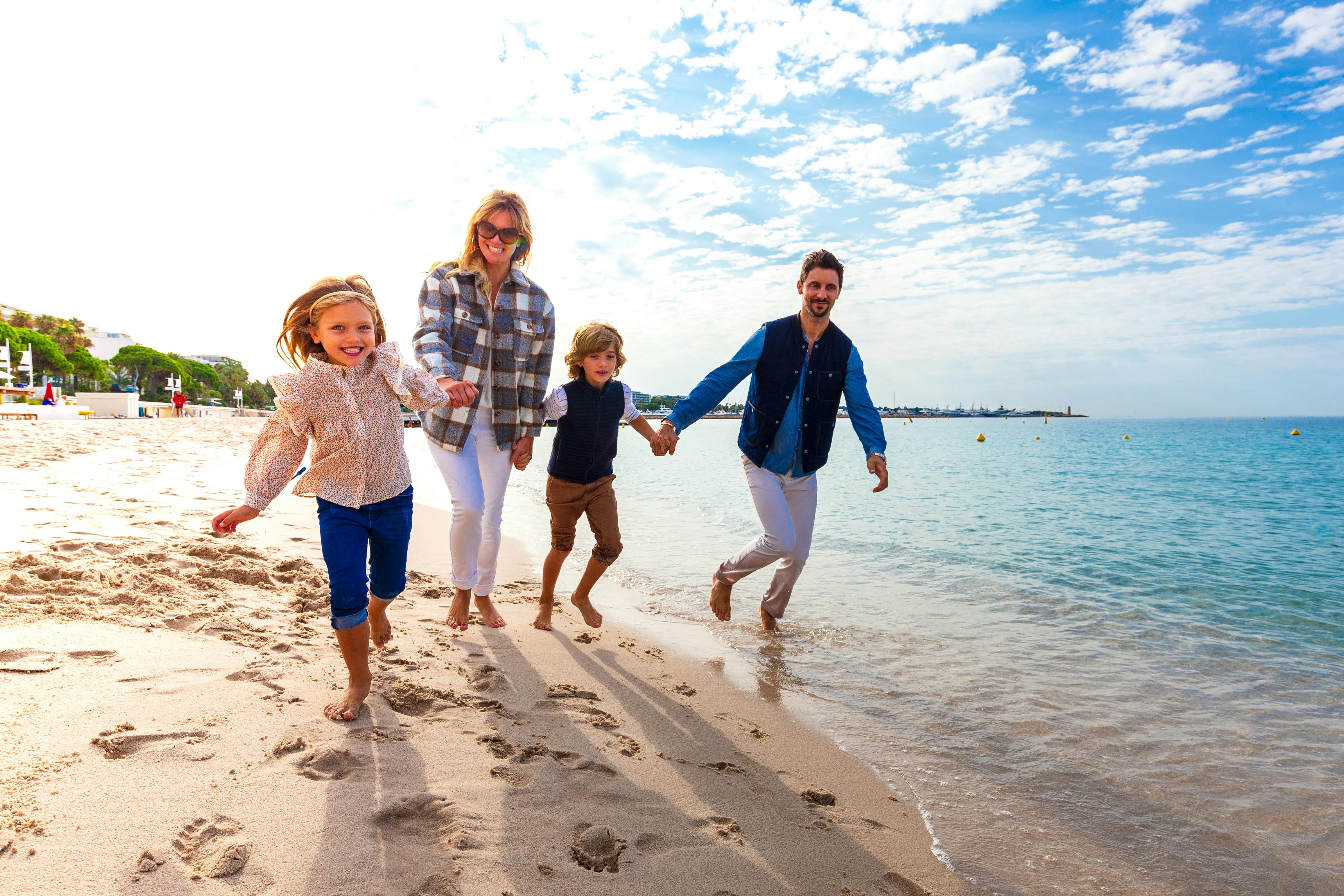 Una família somrient de quatre persones agafades de la mà i caminant per una platja de sorra amb l'oceà i un cel brillant i ennuvolat de fons.
