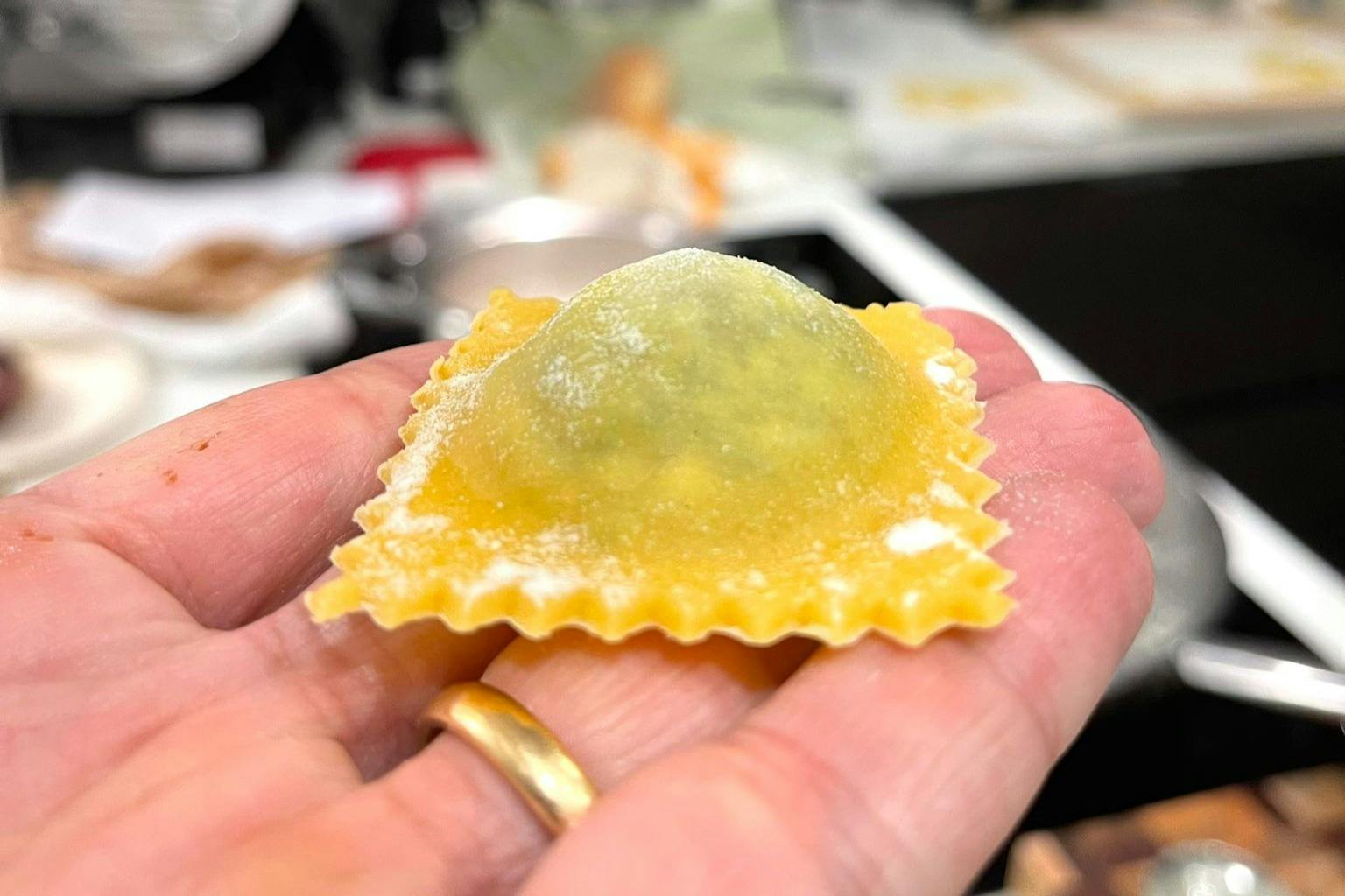 A close-up of a hand holding an uncooked, square ravioli with ruffled edges. The filling inside is visible through the thin pasta.