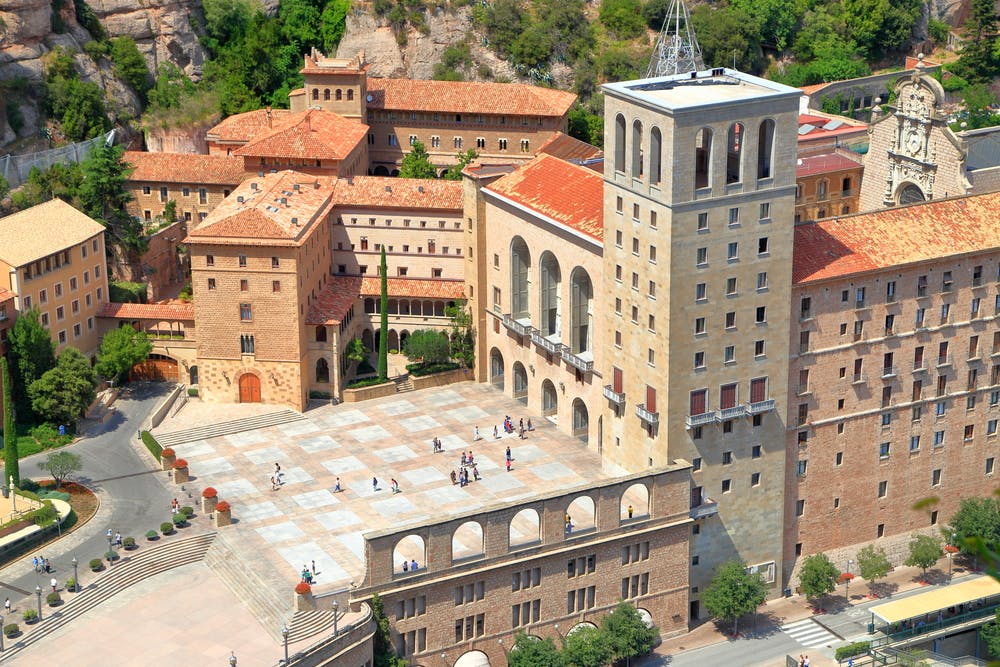 A large courtyard surrounded by historic stone buildings with red-tiled roofs. People are scattered across the courtyard.