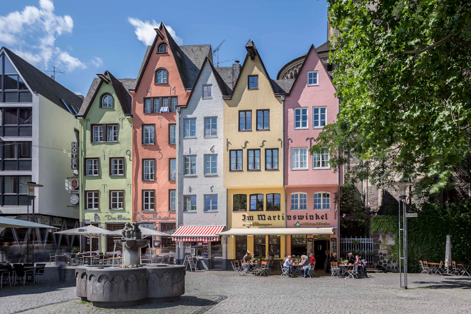 Colorful row of tall, narrow buildings with steep roofs and outdoor seating in front of a restaurant. A fountain is in the foreground.