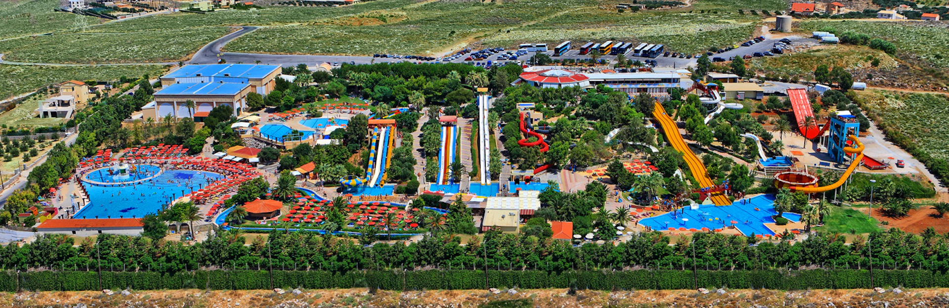 Aerial view of a water park featuring colorful waterslides, pools, surrounding greenery, and nearby buildings and parked vehicles.
