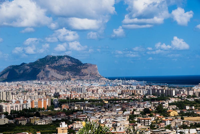 Vue d'un paysage urbain avec de nombreux bâtiments, une montagne en arrière-plan et la mer sous un ciel partiellement nuageux.