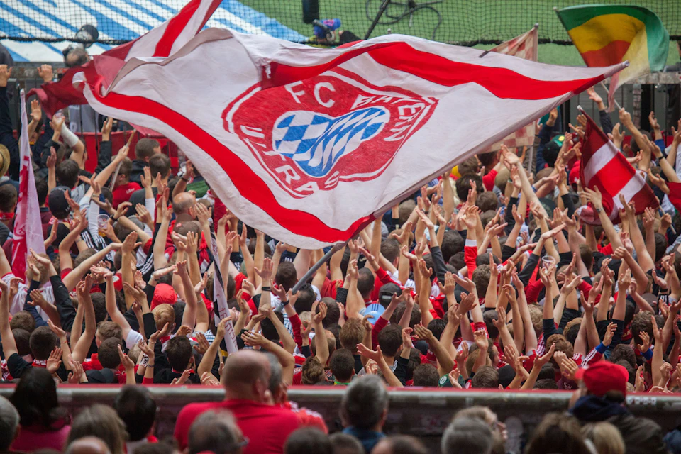 Fans wearing red cheer in a stadium, waving a large white and red flag with "FC Bayern" written on it.