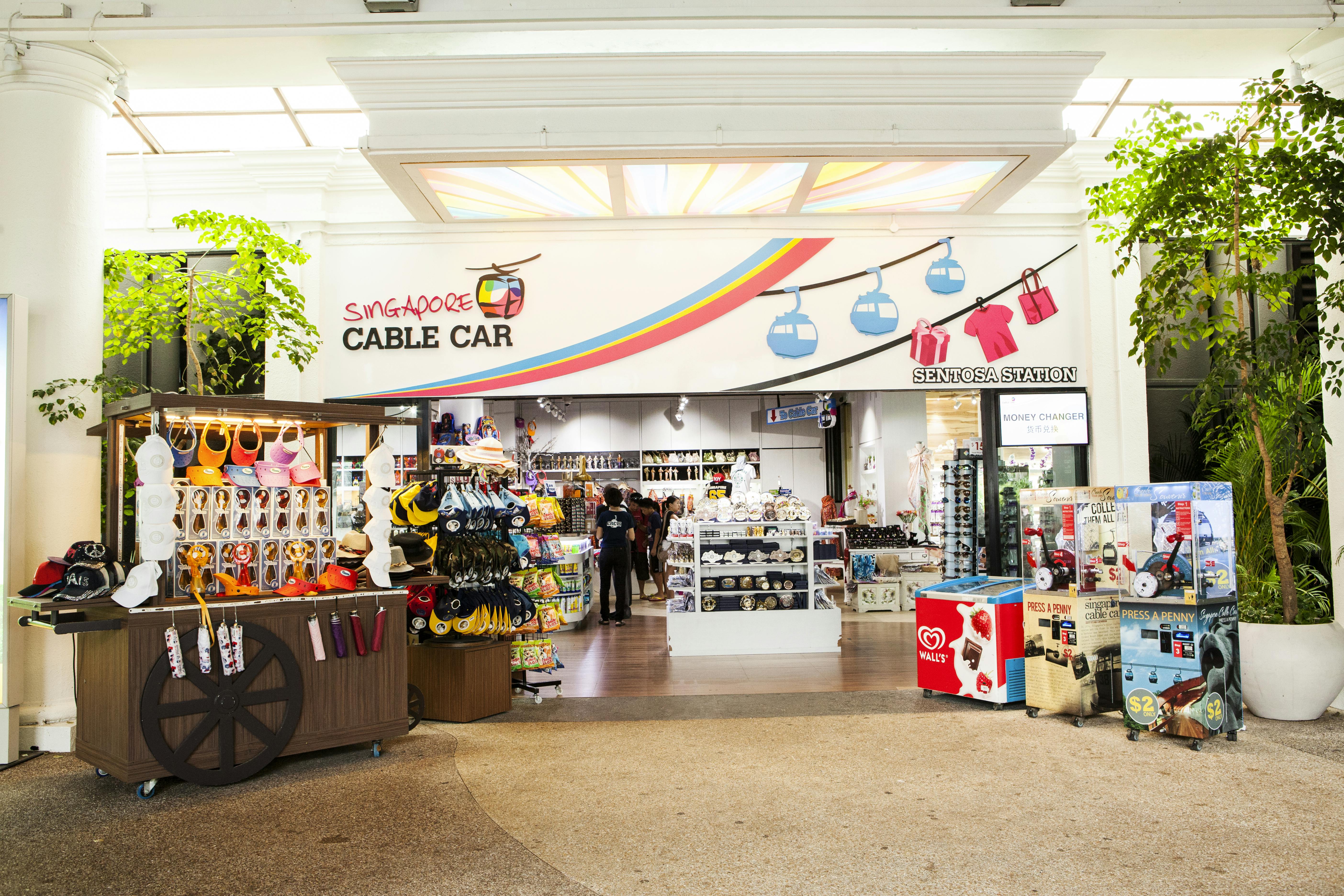 L&#39;entrée d&#39;une boutique de souvenirs intitulée &#34;Singapore Cable Car&#34;, où sont exposés des chapeaux, des jouets et des vêtements. Deux personnes se tiennent à l&#39;intérieur.