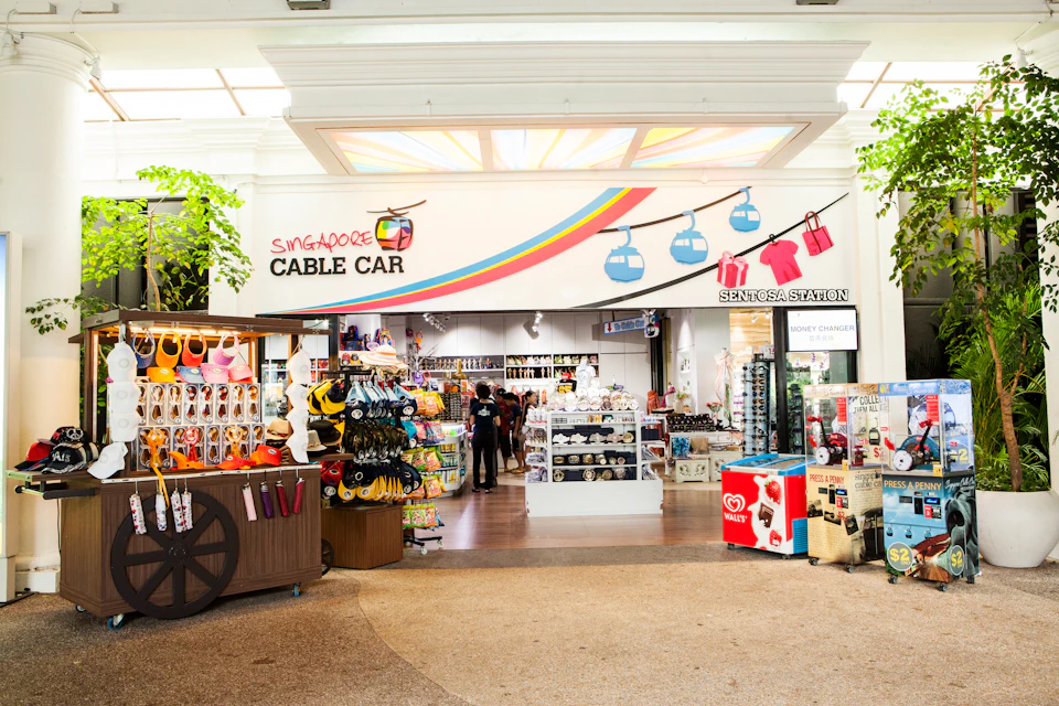 A gift shop entrance labeled "Singapore Cable Car," displaying souvenirs such as hats, toys, and clothes. Two people stand inside.