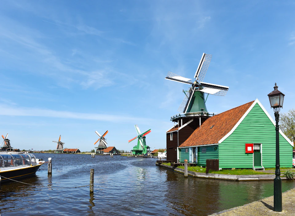 Several traditional windmills positioned alongside a canal with a green, red-roofed building in the foreground. Blue sky overhead.
