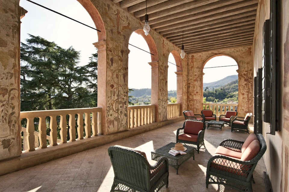 A covered outdoor balcony with decorated arches, wicker chairs, and a table with books overlooking trees and hills.