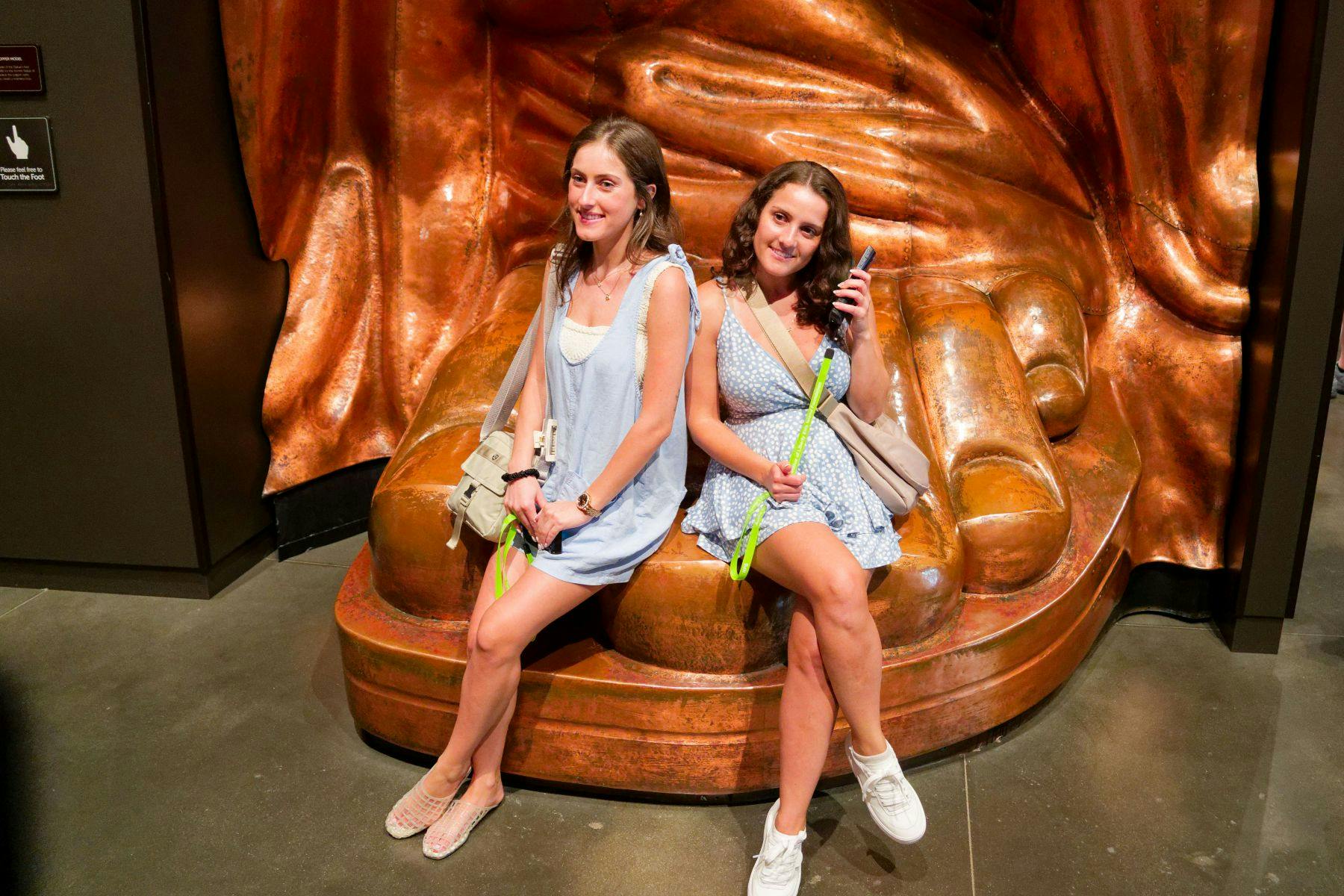 Twee vrouwen zitten glimlachend op de hand van een groot bronzen beeld. Ze zijn casual gekleed en houden groene touwen vast.