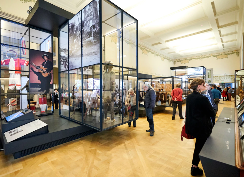 People explore a museum exhibit with glass display cases showcasing historical artifacts and photographs.