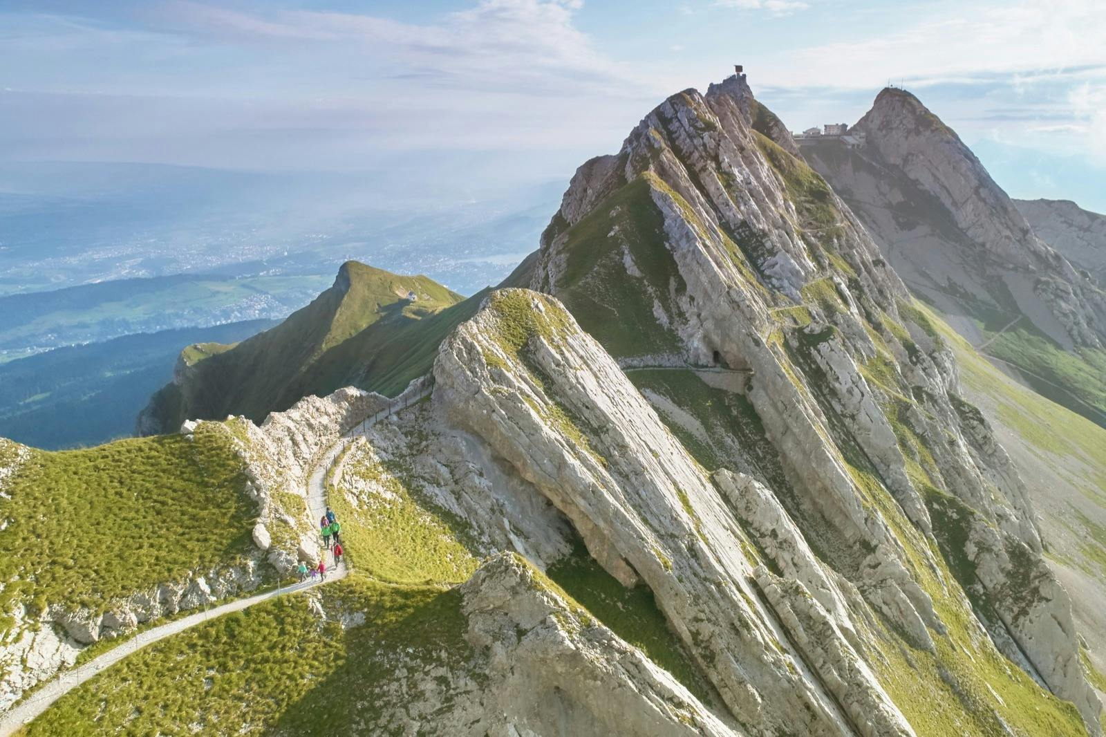 Οι πεζοπόροι περπατούν σε ένα στενό ορεινό μονοπάτι με απότομες, βραχώδεις κορυφές κοντά και ένα τεράστιο τοπίο στο βάθος.