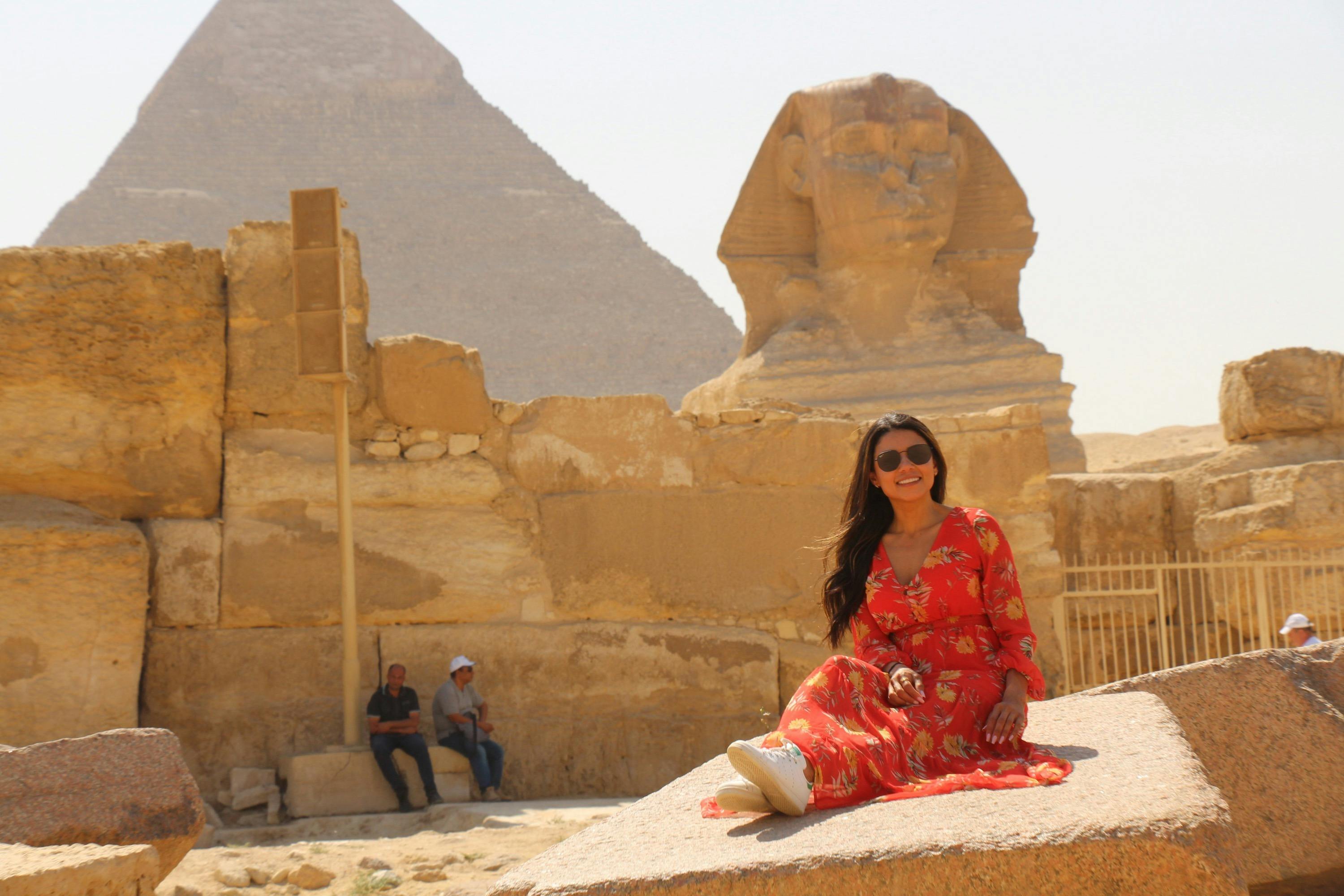 A woman in a red dress sitting on a stone structure in front of the Sphinx in Egypt with a pyramid in the background.