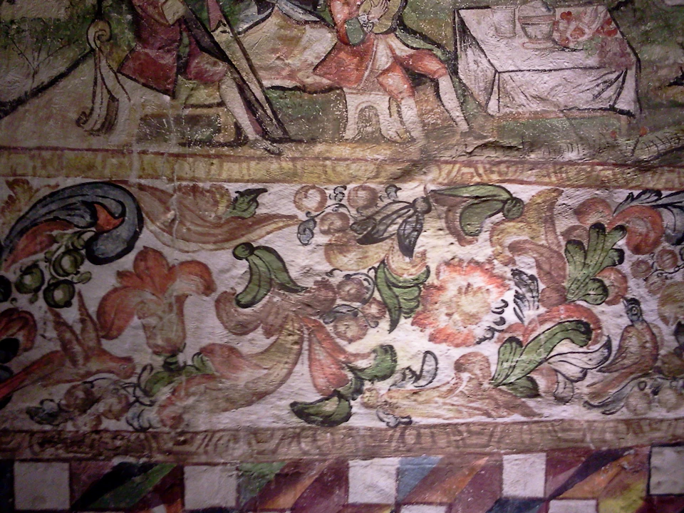 A close-up of a colorful, detailed fresco showing floral patterns, a geometric border, and figures around a table.