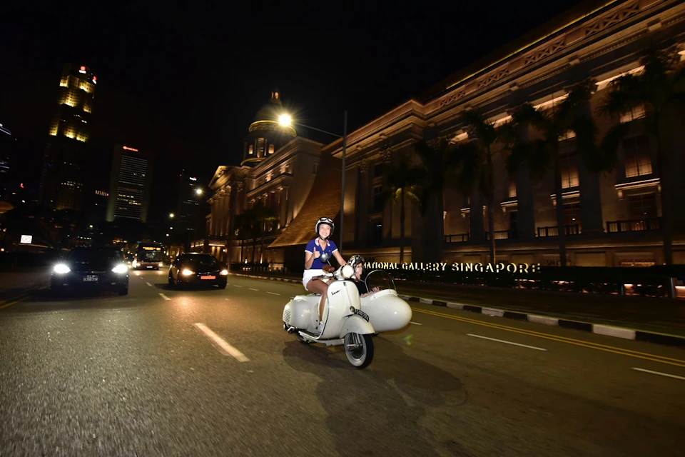 Person riding a white scooter at night on a street with the National Gallery Singapore illuminated in the background.