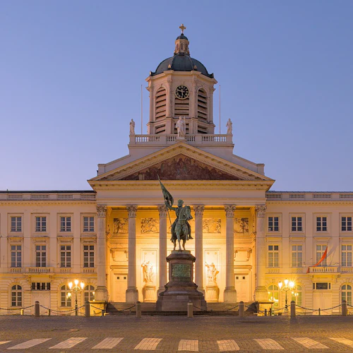 Upplyst neoklassisk byggnad med en central kupol och klocktorn, flankerad av pelare och en staty på hästryggen framför.