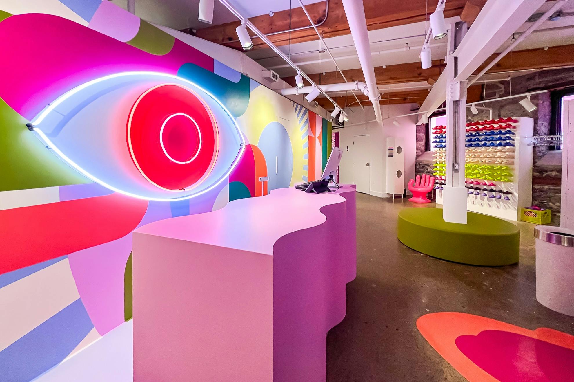 Φωτεινά χρωματισμένο εσωτερικό με ροζ κυματιστό γραφείο, σχεδιασμό ματιών νέον και ζωντανή διακόσμηση, με μοντέρνα έπιπλα και οθόνες.