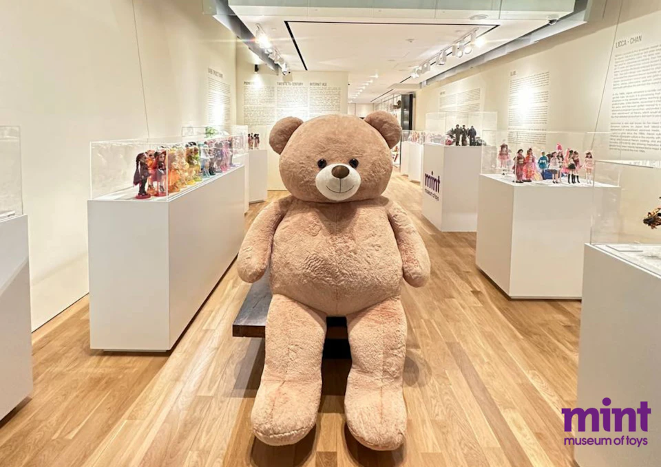 Large tan teddy bear seated on a bench in a museum-style display area filled with glass cases showcasing various dolls.