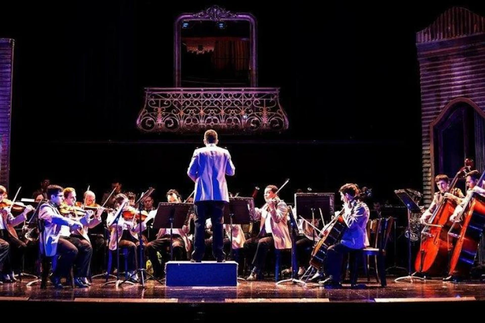 An orchestra performing on stage with a conductor in the foreground, viewed from behind, under ornate balcony railings.