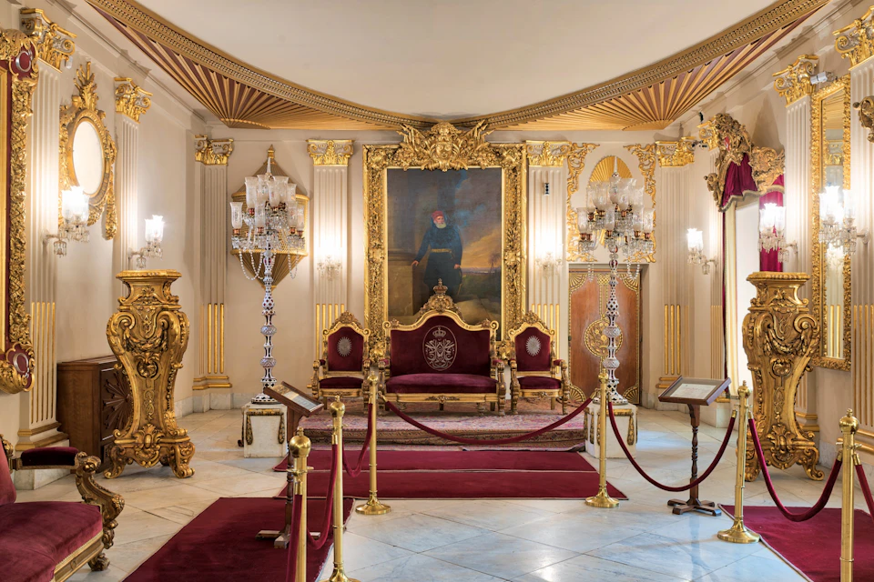 Ornate room with red velvet furniture, gold accents, chandeliers, and a large portrait above a throne, roped off for viewing.