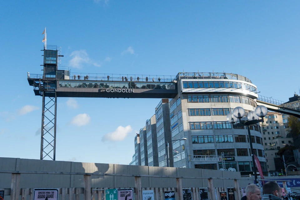 A modern building with a glass walkway labeled "Gondolen," under a clear blue sky, with people visible on the walkway.