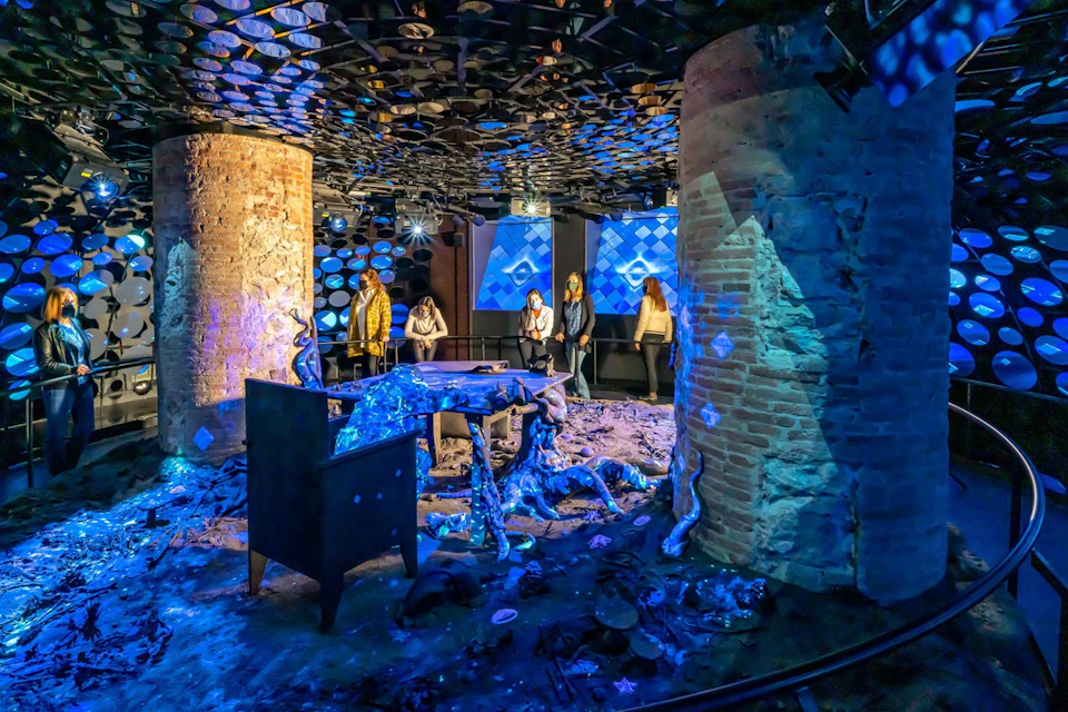 People observe illuminated abstract art in a room with brick columns and reflective ceiling decorations.