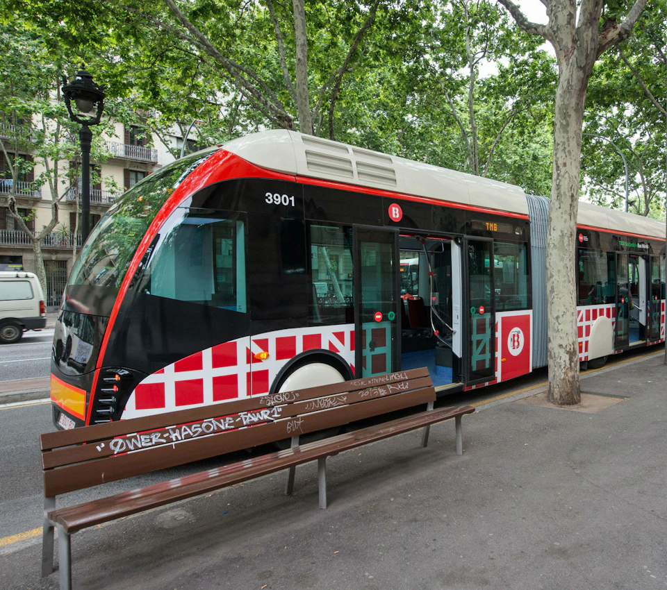 A modern tram with a red and black design parked near benches and trees on a city street.