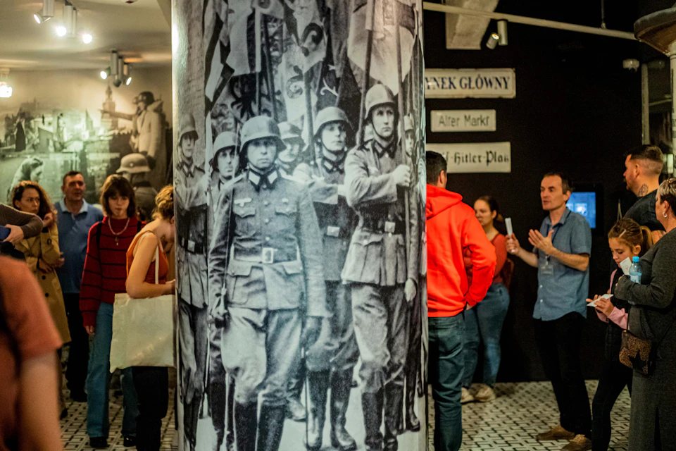 People observing a large photograph of WWII soldiers in a museum exhibit, with street sign replicas on the wall.