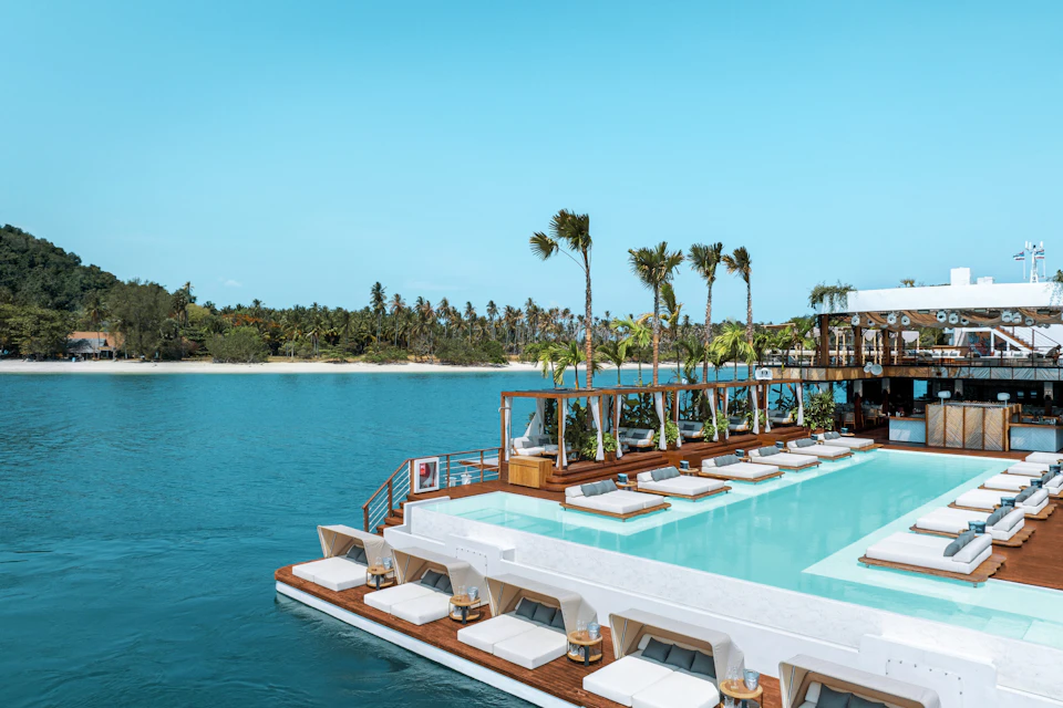 Poolside lounge area on a dock over clear blue water. White lounge chairs and palm trees with an island visible in the background.