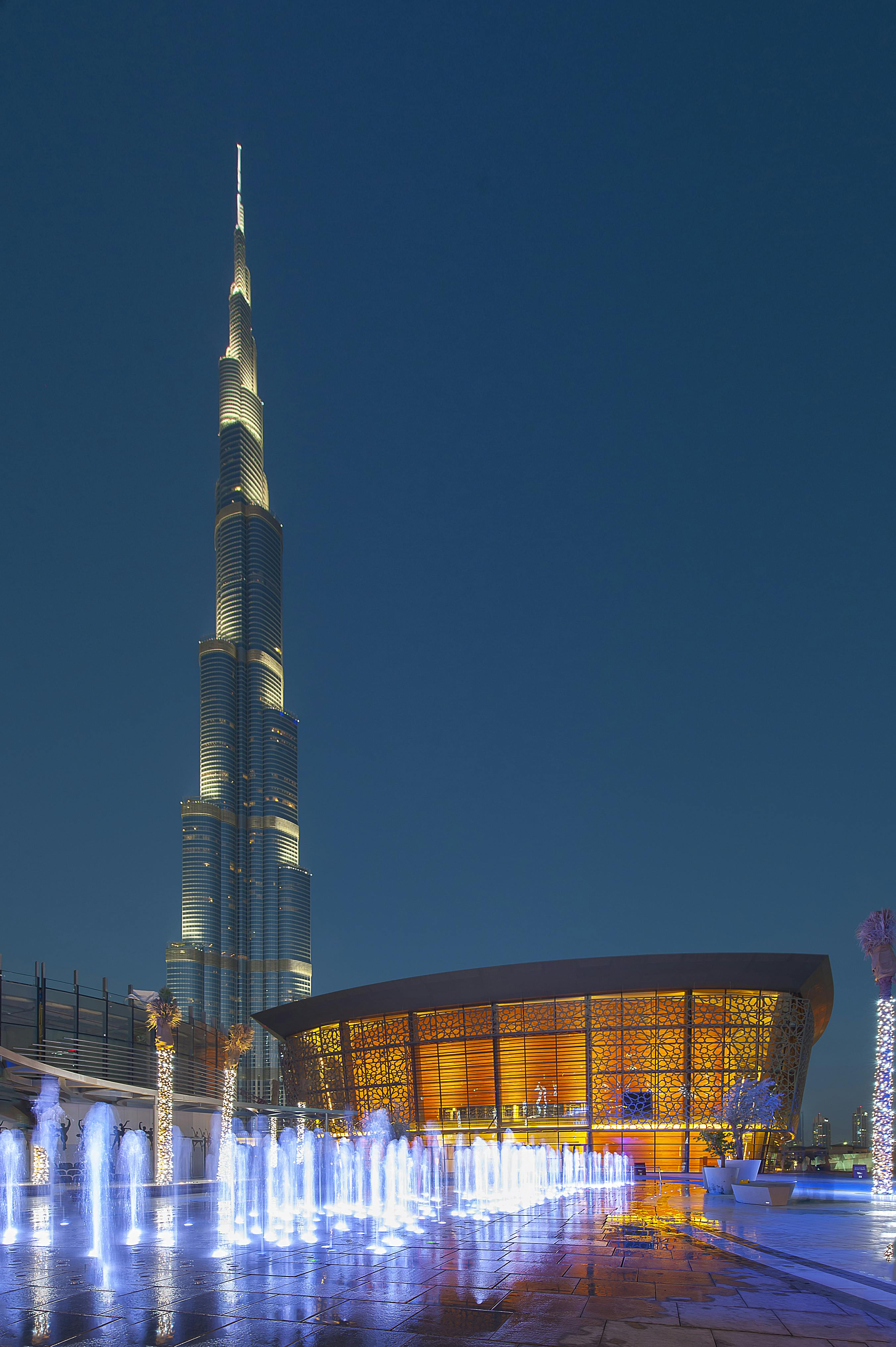 Tall, illuminated skyscraper beside a curved building with lit windows against a twilight sky.