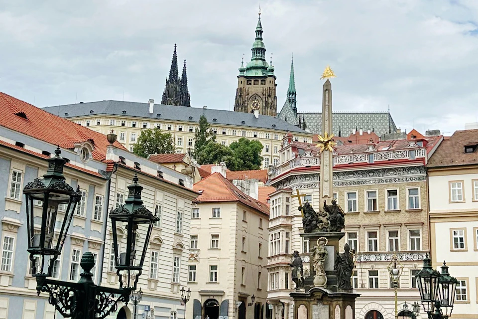 A town square featuring an ornate column with statues, surrounded by historic buildings and a cathedral in the background.