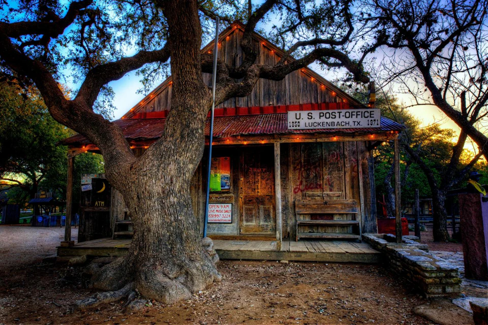 Rustic wooden post office with a large tree in front, located in Luckenbach, Texas. The building has weathered wood and a porch.