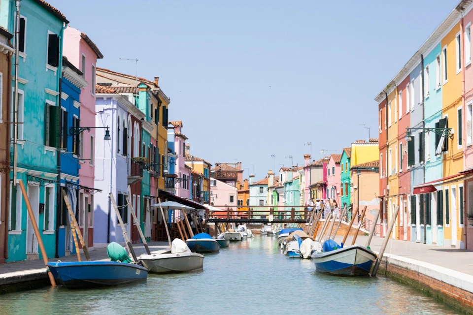 Colorful buildings line a canal with boats docked alongside on a clear day. A small bridge spans the canal in the background.