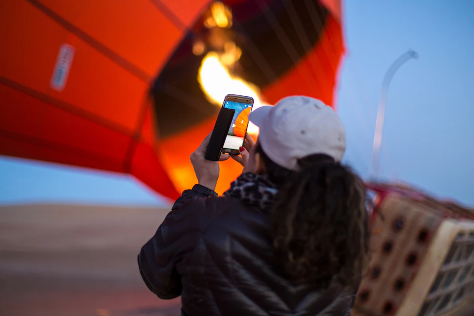 A person in a white cap photographs an inflating hot air balloon with their phone.