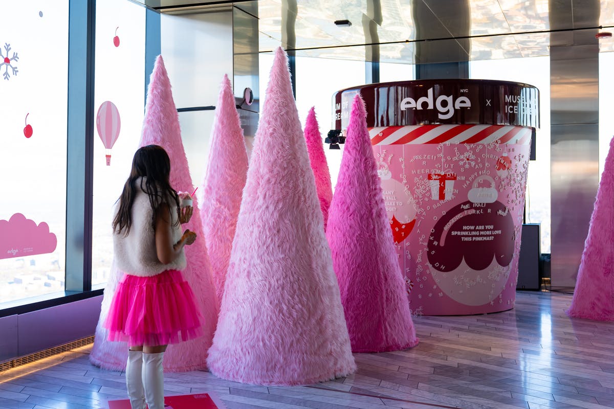 Osoba v růžovém a bílém oblečení stojí v blízkosti vysokých růžových fuzzy kuželovitých dekorací v moderním vnitřním prostoru.