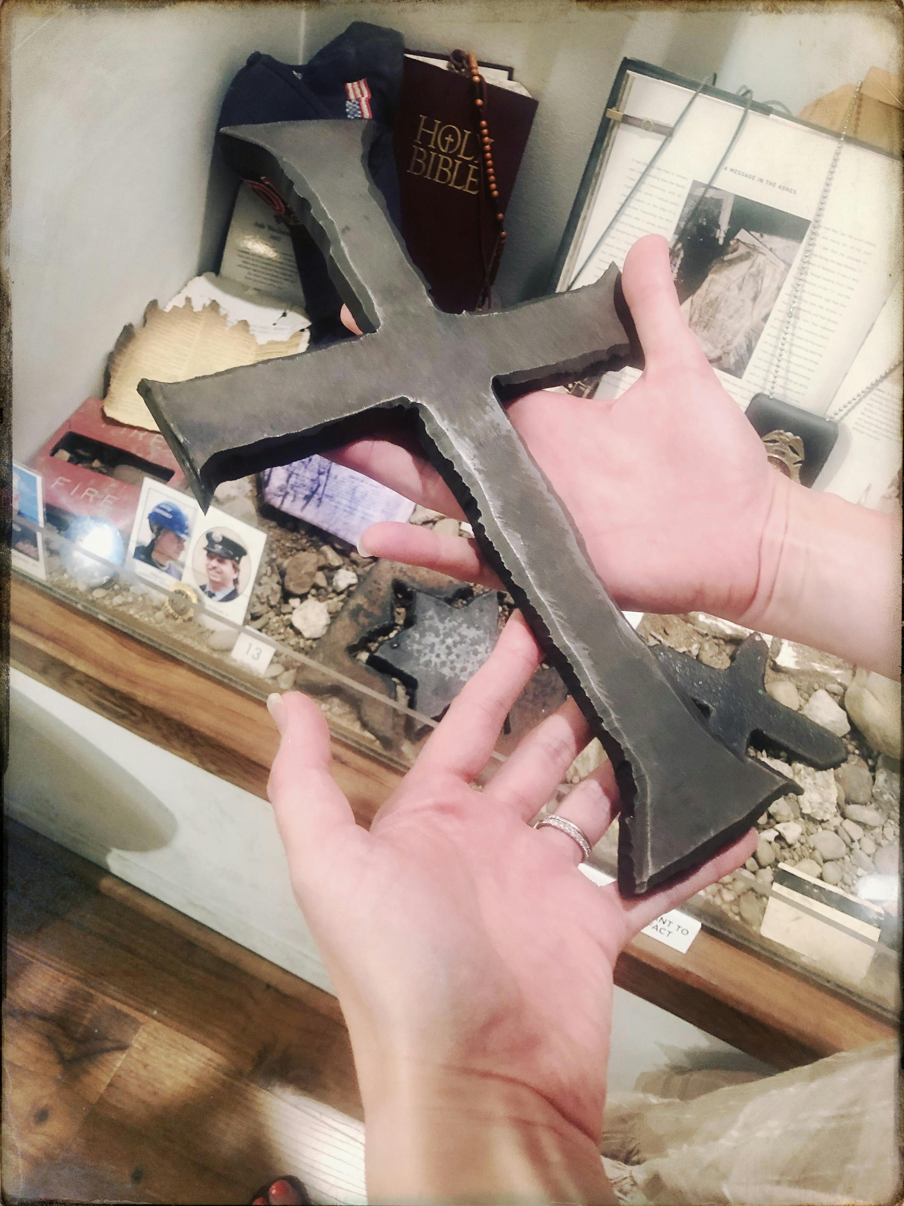 Two hands holding a large black cross, with historical artifacts displayed in the background, including photos and a book.