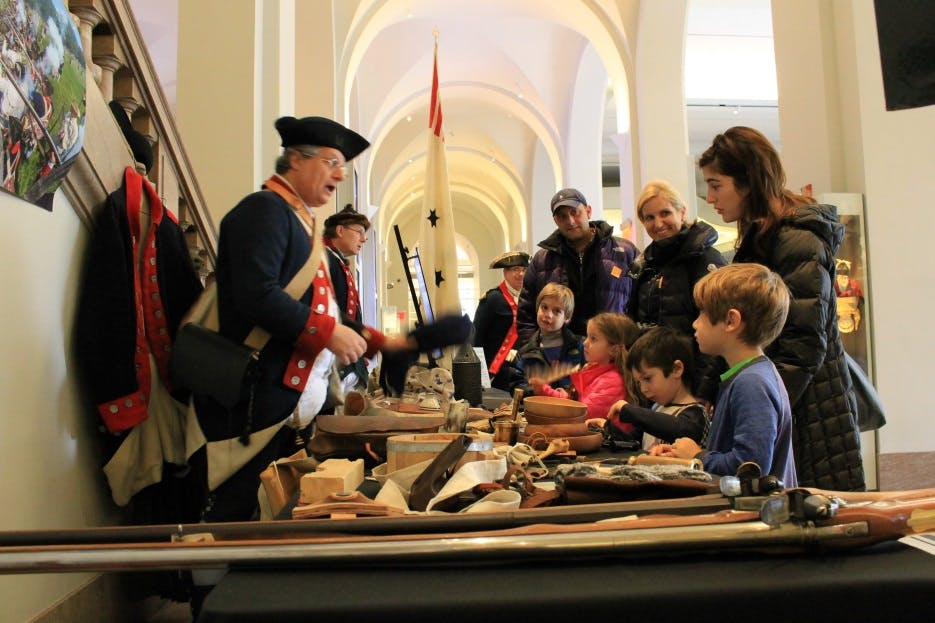 Οι επισκέπτες συγκεντρώνονται γύρω από ένα τραπέζι με ιστορικά αντικείμενα, ακούγοντας έναν παρουσιαστή με κοστούμι εποχής. Τα παιδιά και οι ενήλικες συμμετέχουν ενεργά.