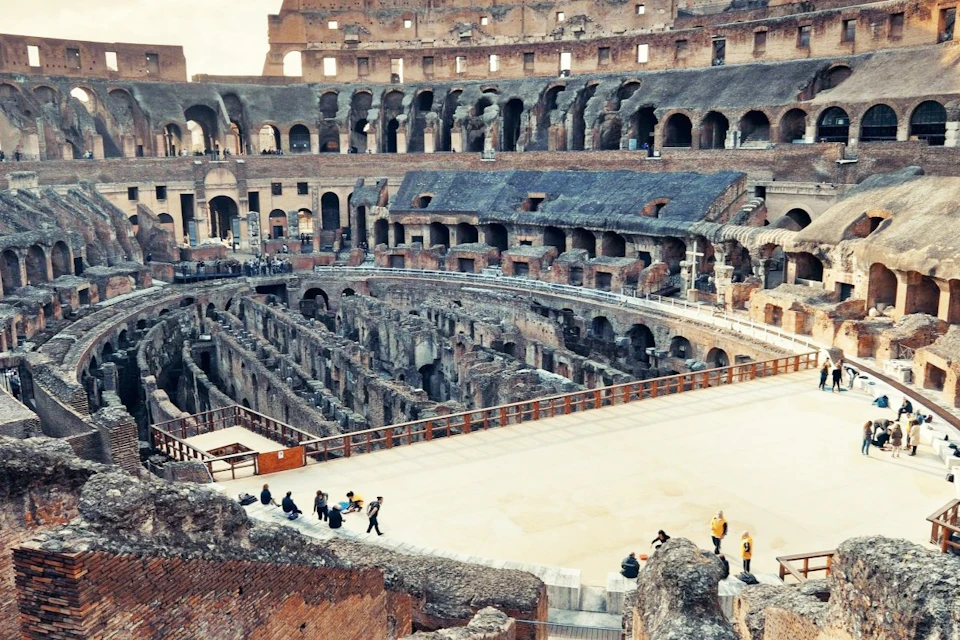 View of the interior of the Colosseum in Rome, showing its ancient stone structure and tourists exploring the site.