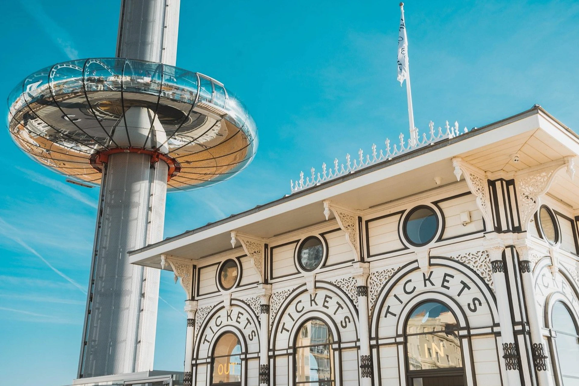 A ticket booth in ornate style beside a modern tower with a circular observation pod, under a clear blue sky.