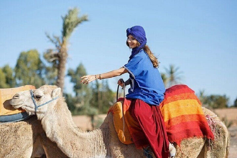 Una persona que porta un mocador blau i una faldilla vermella munta un camell cobert amb mantes de colors en un paisatge desert ple de palmeres.