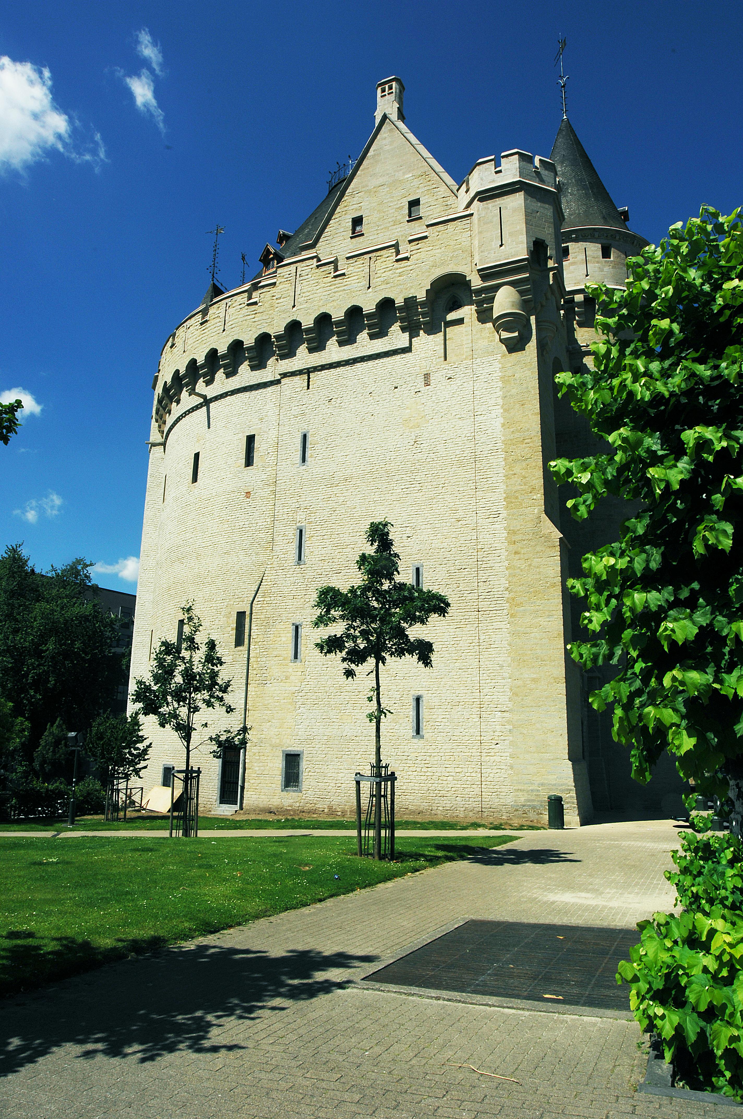 Tour historique en pierre avec des murs arrondis et des tourelles, entourée d'une pelouse, d'arbres et d'un chemin pavé sous un ciel bleu clair.