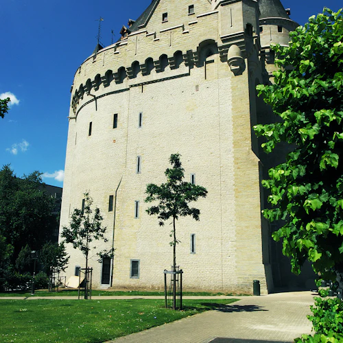 Historiskt stentorn med rundade murar och torn, omgivet av en gräsyta, träd och en kullerstensstig under en klarblå himmel.