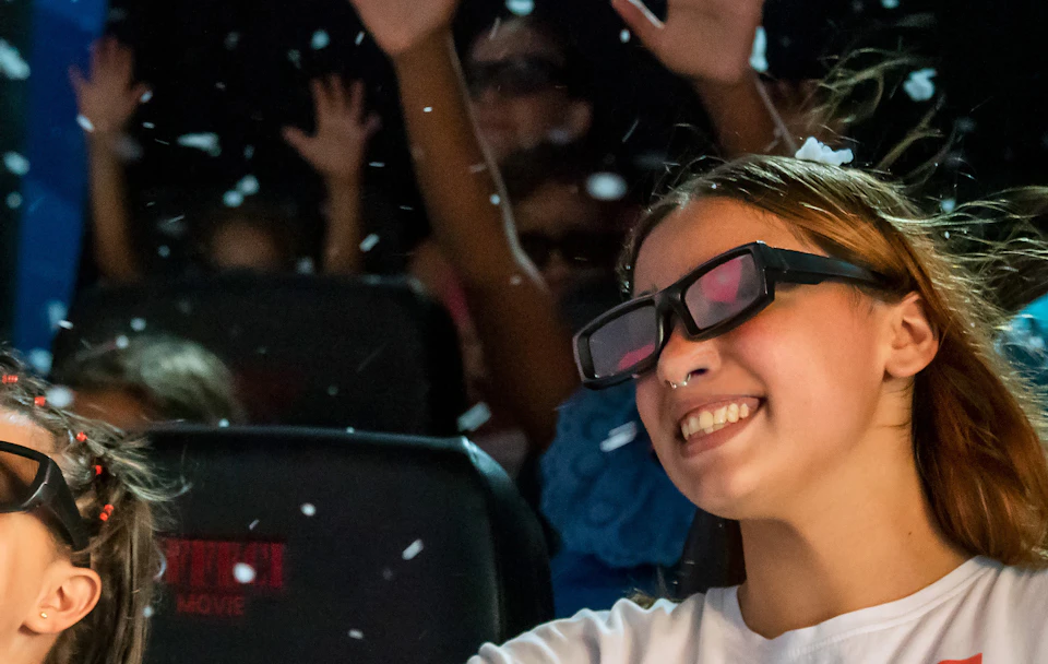 Children and adults wearing 3D glasses sit in a theatre, smiling and raising hands, with confetti or snow falling around them.